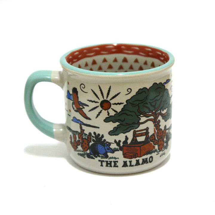 The Alamo Feeling Folky Mug is a ceramic mug with a light blue handle, vibrant illustrations of the Alamo, trees, sun, birds, cactus, THE ALAMO text, and a red geometric pattern inside—perfect for celebrating Texas history.