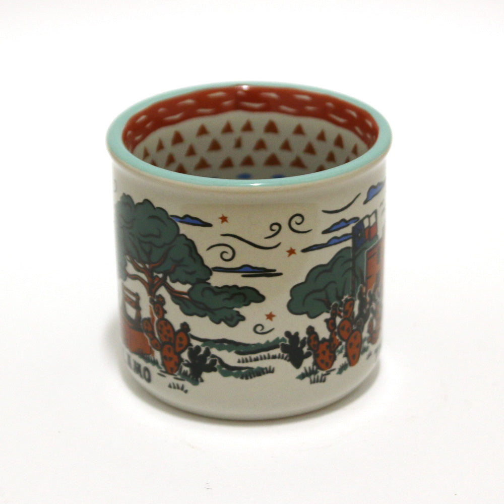 The Alamo Feeling Folky Mug is a white ceramic mug featuring colorful illustrations of trees, cacti, clouds, stars, and a Texas-inspired building. The inside rim displays a red triangular pattern.
