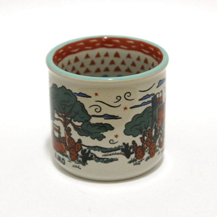 The Alamo Feeling Folky Mug is a white ceramic mug featuring colorful illustrations of trees, cacti, clouds, stars, and a Texas-inspired building. The inside rim displays a red triangular pattern.