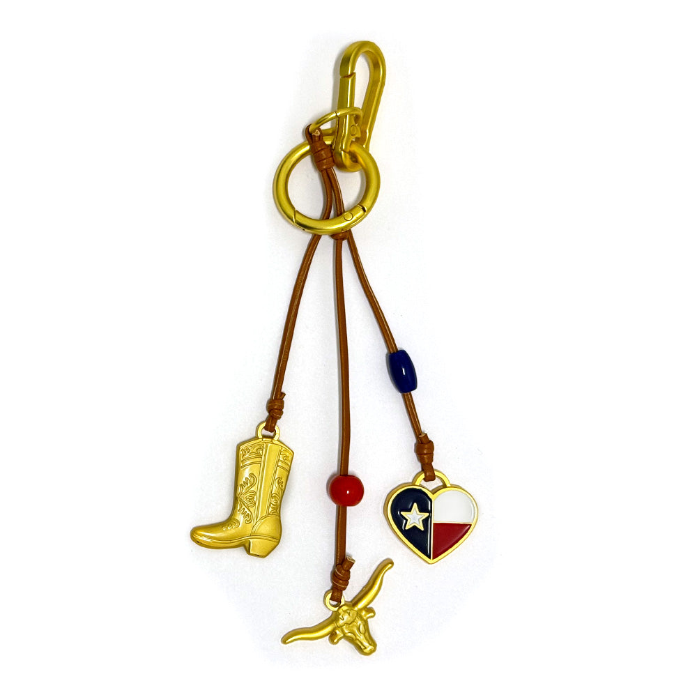 The Texas Icon Trio Bag Clip features gold hardware, two brown leather cords—one with a cowboy boot charm and red bead, the other with a heart-shaped Texas flag charm, blue bead, and gold longhorn. A perfect gift for Texan pride!.