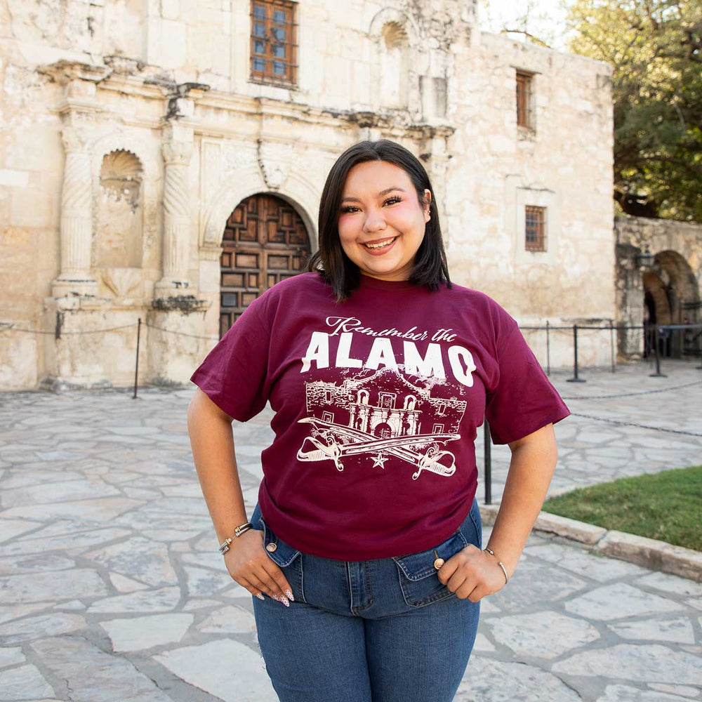 A woman wearing a maroon 100% cotton Remember the Alamo T-Shirt featuring a graphic of the Alamo and crossed swords, standing in front of the Alamo building.
