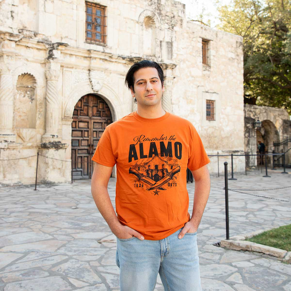 A man wearing a bright orange graphic t-shirt with 'Remember the ALAMO 1843-1973' printed on the front, standing outdoors in front of a historic stone building.