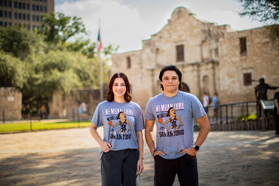 Two people stand smiling in front of the Alamo in San Antonio, Texas, both wearing matching San Antonio Spurs Victor Wembanyama T-Shirts featuring a cartoon basketball player and the text “Wembanyama San Antonio.” Their shirts celebrate the excitement surrounding the Spurs’ rising star.