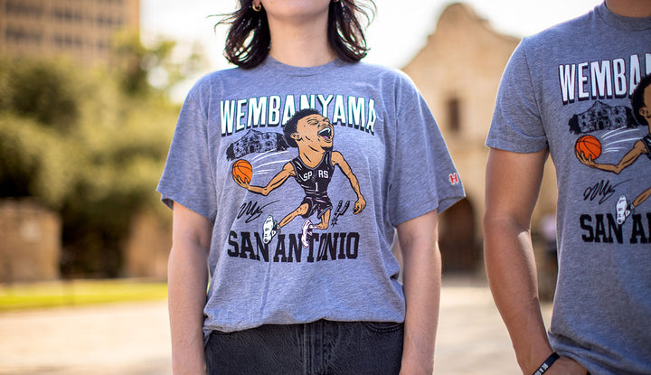 A person wearing a gray San Antonio Spurs Victor Wembanyama T-Shirt featuring a cartoon basketball player holds another person’s arm; both are outside with blurred historic buildings in the background.