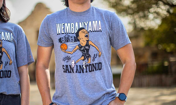 A person is standing outdoors wearing the San Antonio Spurs Victor Wembanyama T-Shirt, which is gray and features a cartoon basketball player holding a ball, along with the text “Wembanyama San Antonio” and the Spurs logo.