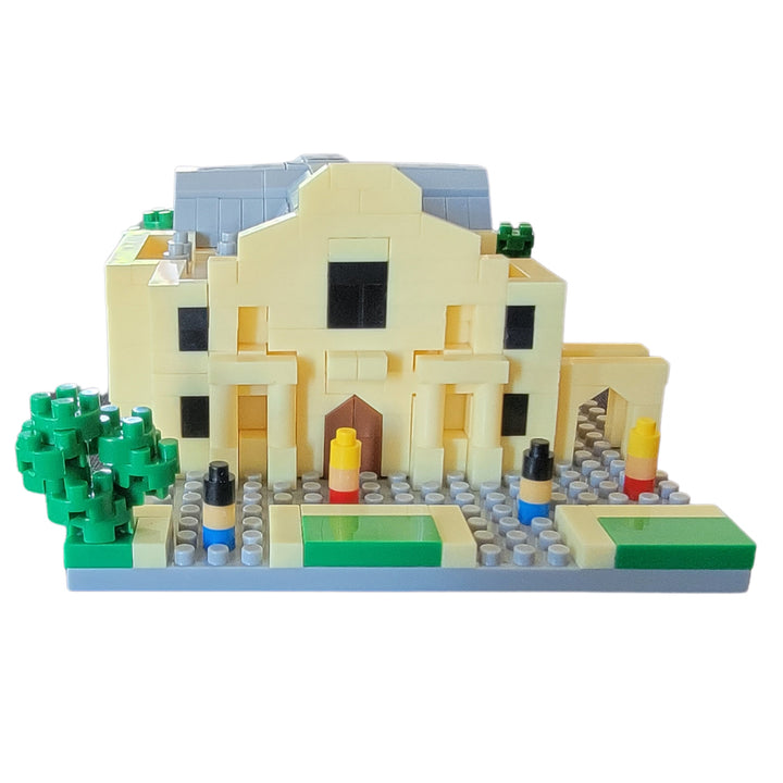 The Mini Alamo Building Block Set features a yellow building with a gray roof, central entrance, windows, trees, and small pillars on a rectangular base. Includes assembly instructions for easy construction.
